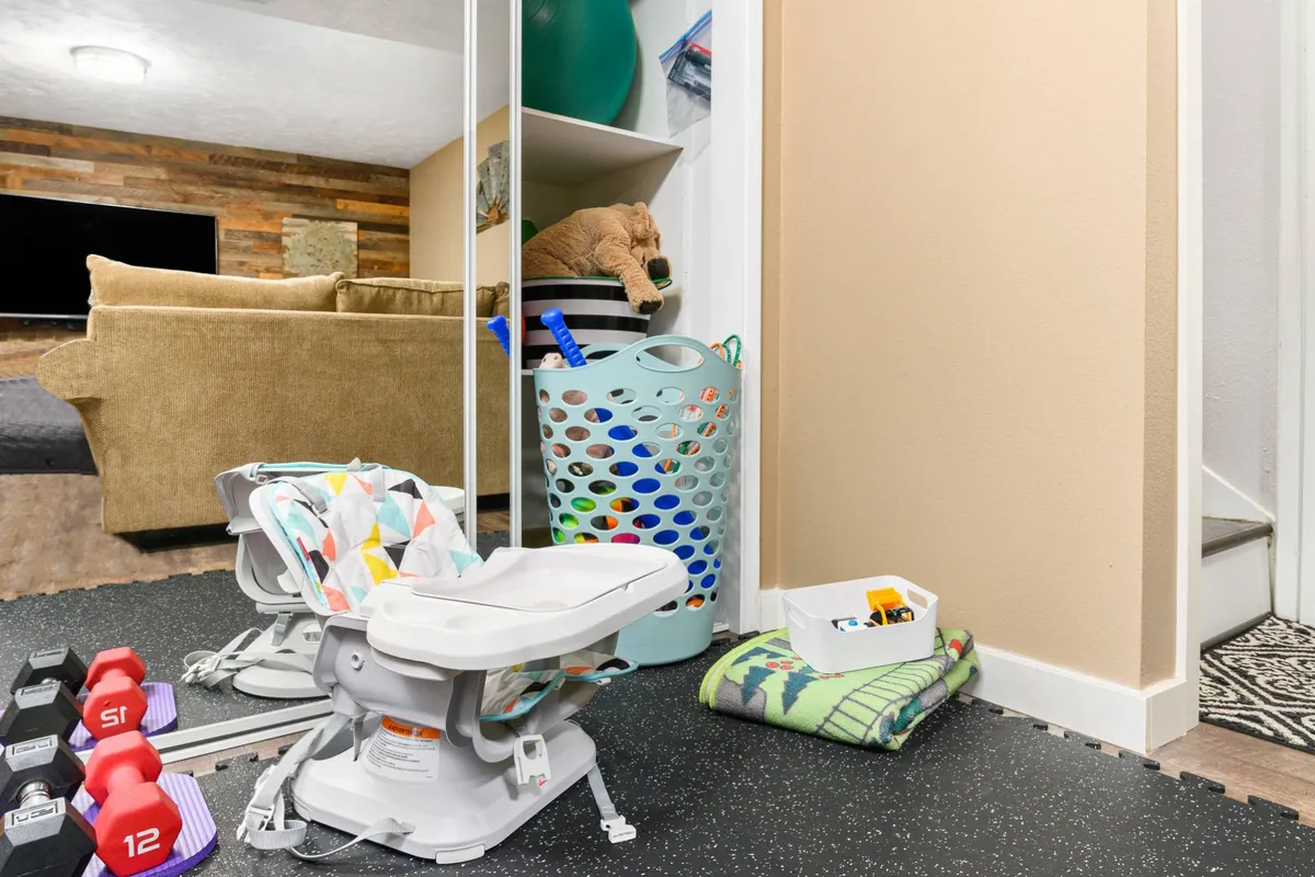 This image features a cozy corner with a baby chair, dumbbells, a storage basket, and a couch, highlighting the multi-functional aspect of the space. The presence of gym equipment signals an area dedicated to fitness, while the soft furnishings emphasize comfort and relaxation.