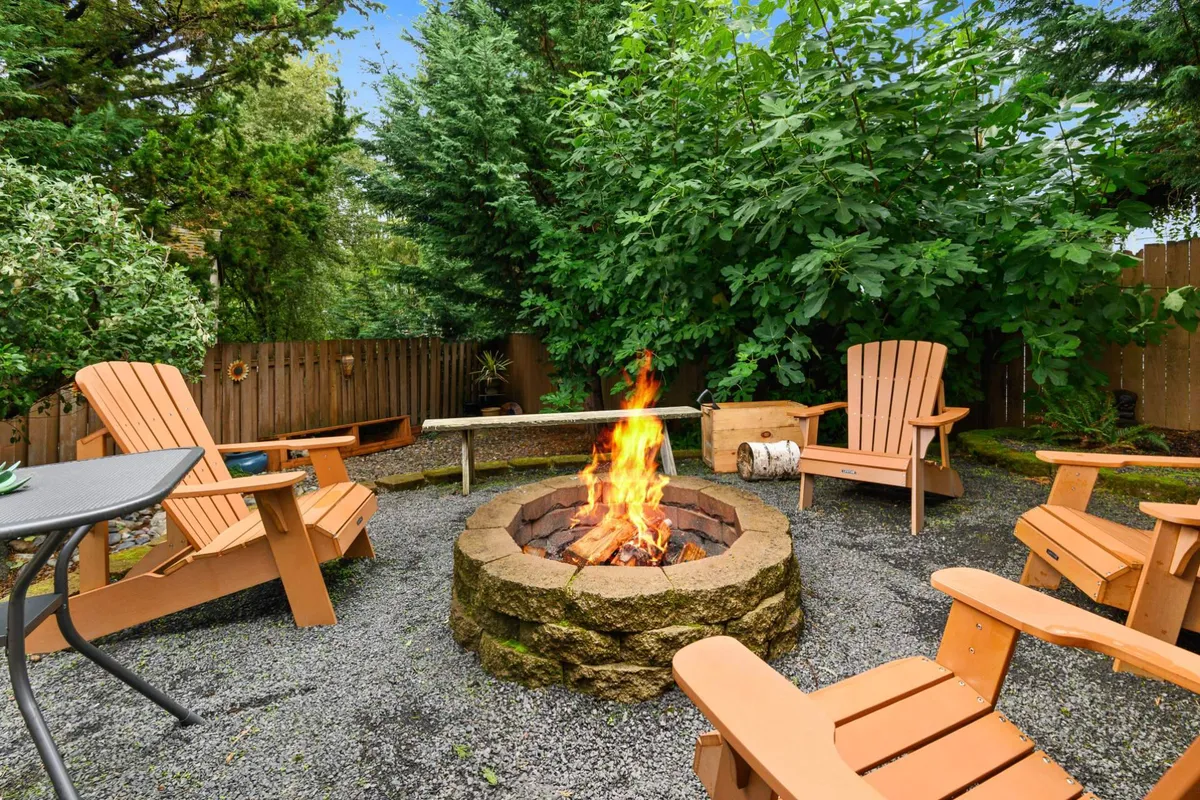 A welcoming backyard scene featuring a central fire pit surrounded by wooden chairs, set amidst abundant greenery. The atmosphere evokes warmth, relaxation, and the inviting glow of an evening gathering.