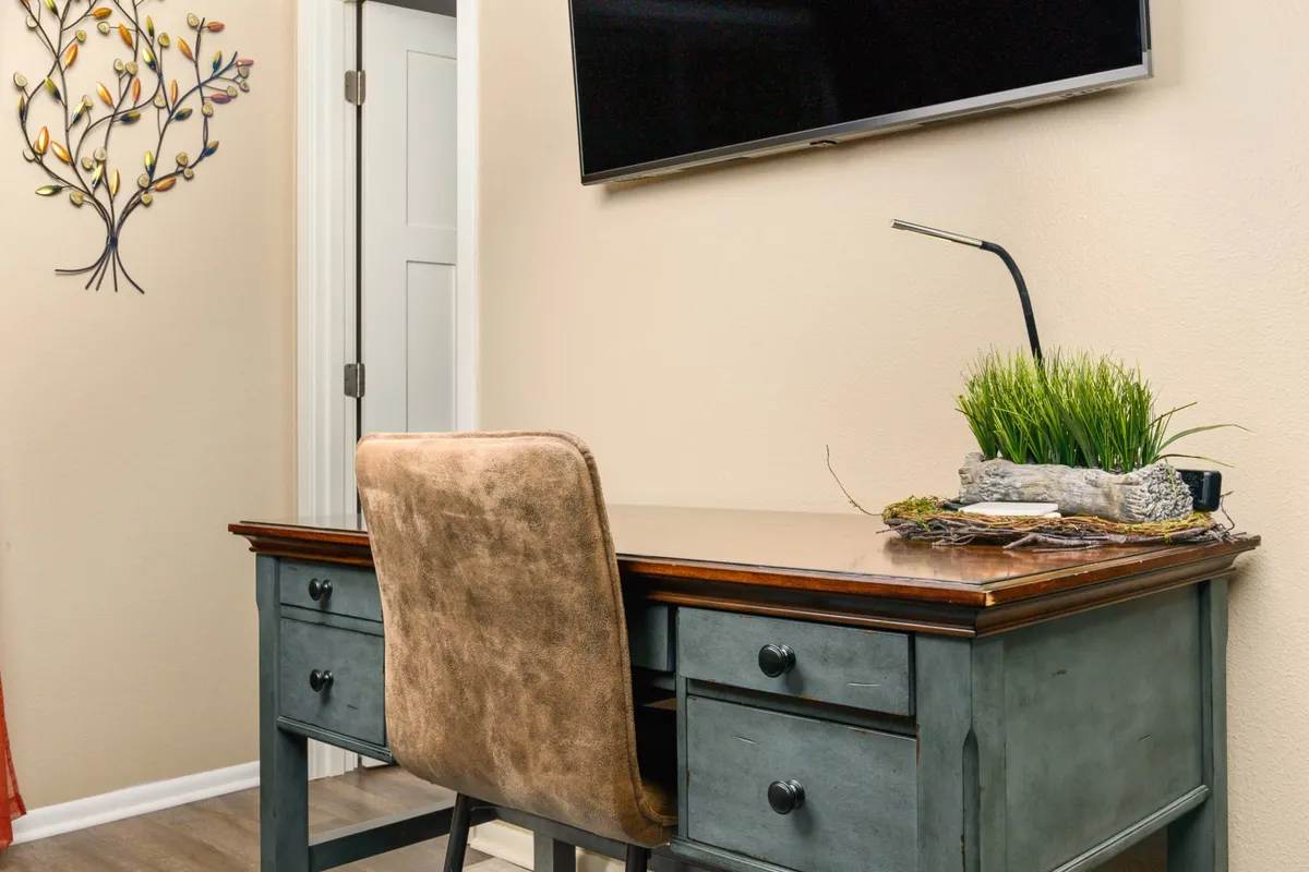 A serene, dedicated workspace featuring a wooden desk, ergonomic chair, potted plant, and a TV mounted on the wall. The setting is ideal for focused work, study, or enjoying entertainment, blending productivity with relaxation.