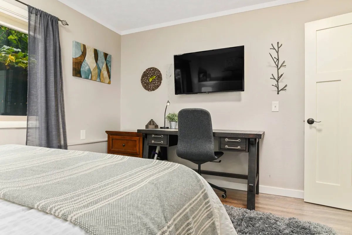 A secondary bedroom featuring a cozy bed with a textured blanket, a convenient desk and chair for versatile use (work or reading), and natural light streaming in. Decorative wall art and a wall-mounted TV add contemporary touches, supporting a multi-functional and welcoming environment.