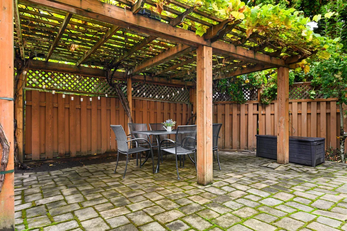 A tranquil outdoor dining area beneath a charming pergola, with a table set for relaxation and potted plants adding to the lush, green ambiance. The pergola is draped with vines, offering both beauty and shade.