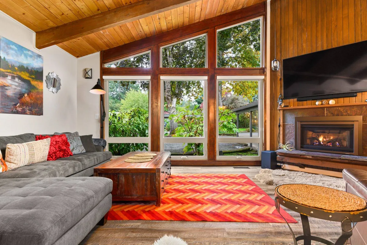The hero image captures a stylish, sunlit living room with a large sectional sofa facing a cozy fireplace. Expansive windows fill the space with natural light and offer lush views of surrounding trees, while a vibrant area rug and wooden coffee table add a touch of mid-century charm. The setting feels warm and inviting, perfect for gathering with loved ones, relaxing with a book, or watching the seasons change from the comfort of home.