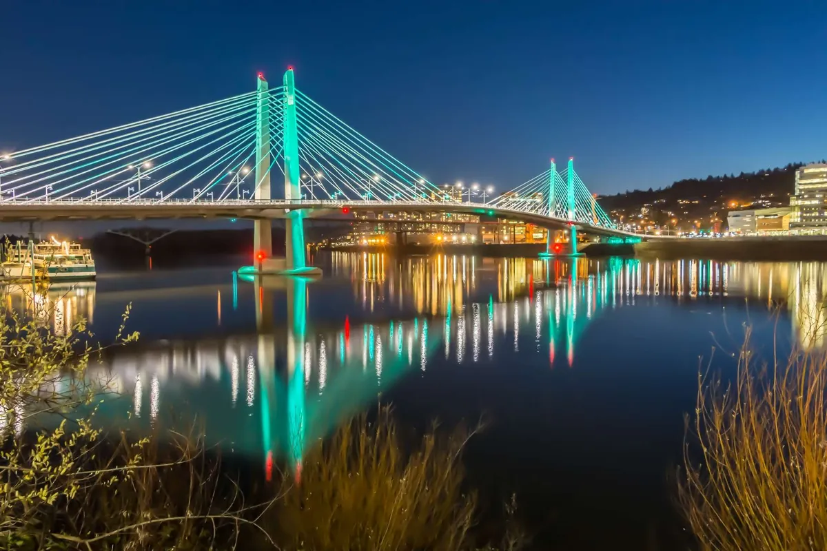 A nighttime view of an illuminated bridge spanning a tranquil river, with city lights glowing in the background and reflections shimmering on the water. The setting suggests a peaceful yet lively environment perfect for unwinding after a busy workday or enjoying a scenic walk.