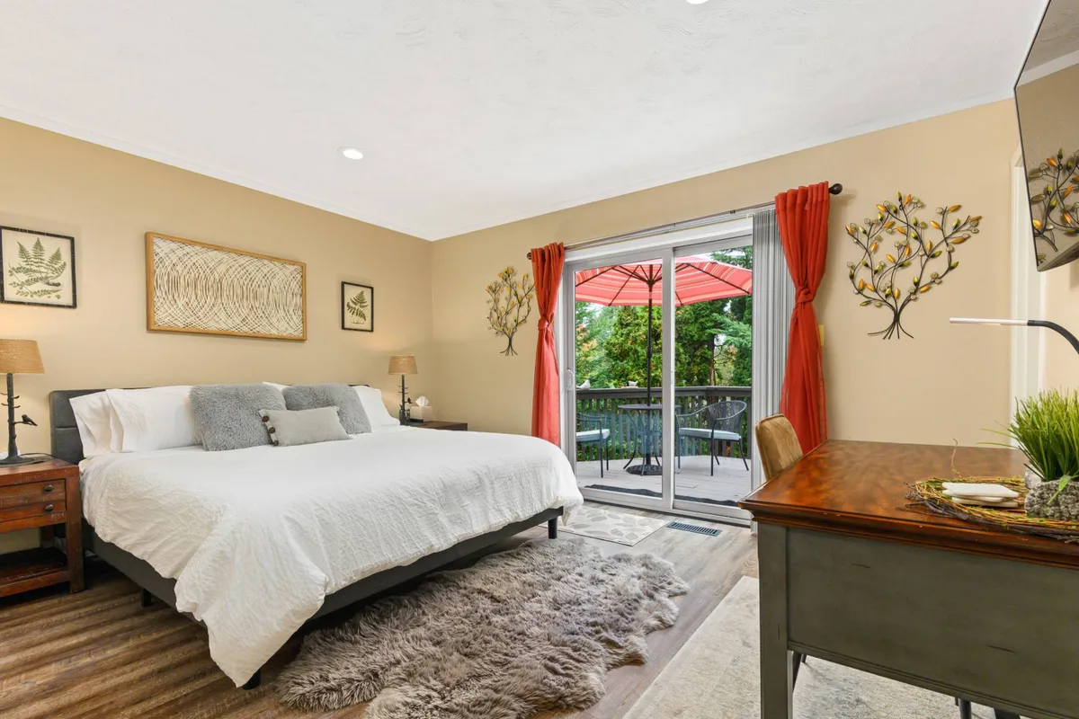 A spacious bedroom with a large bed and soft linens, enhanced by a sliding glass door that floods the room with daylight. Features restful décor, a desk with a potted plant, and decorative wall art, encapsulating tranquility and the connection to the outdoors.