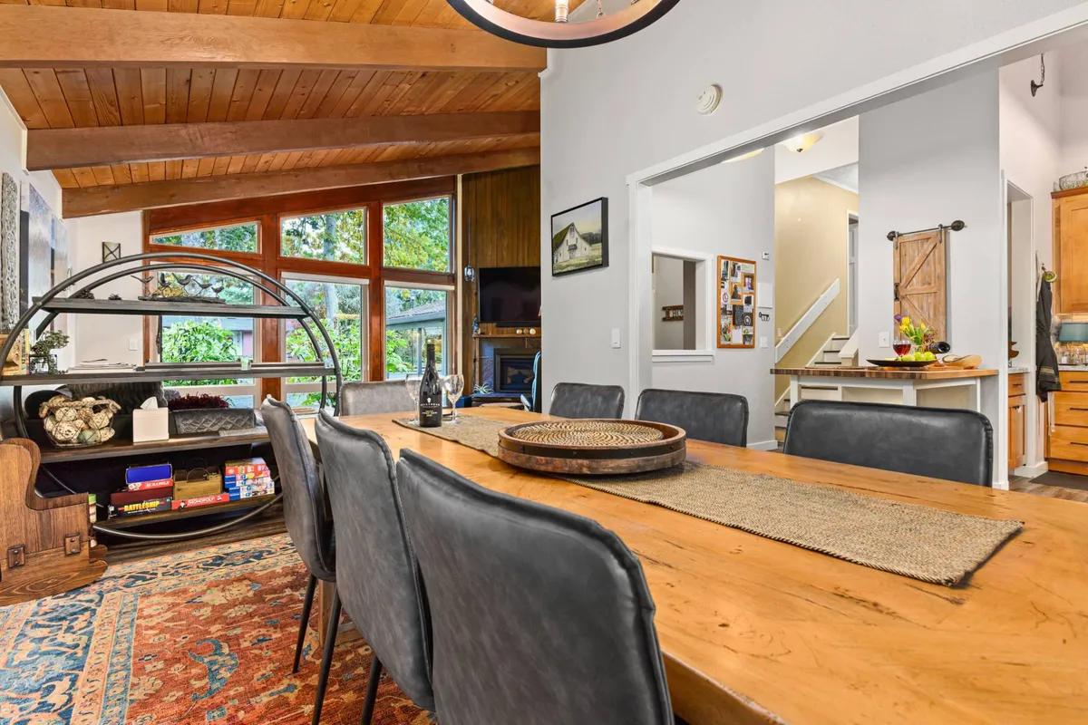 A large wooden dining table beneath a charming chandelier is surrounded by comfortable seating and windows that frame views of the garden. The setting is ready for family meals, entertaining, or casual brunches, with the garden view lending fresh natural light and tranquility.