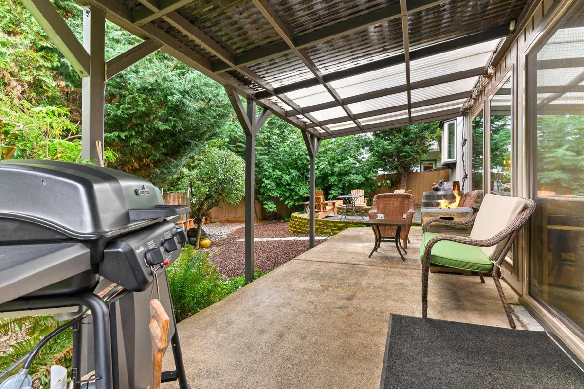 A versatile patio space featuring a grill, comfortable outdoor seating, a fire pit, and a backdrop of vibrant greenery. The setting is ideal for barbecues, drinks by the fire, or simply enjoying the outdoors with friends and family.